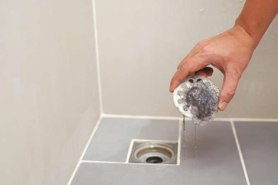 A person removing a clogged hair strainer from a stubborn drain in a bathroom floor.